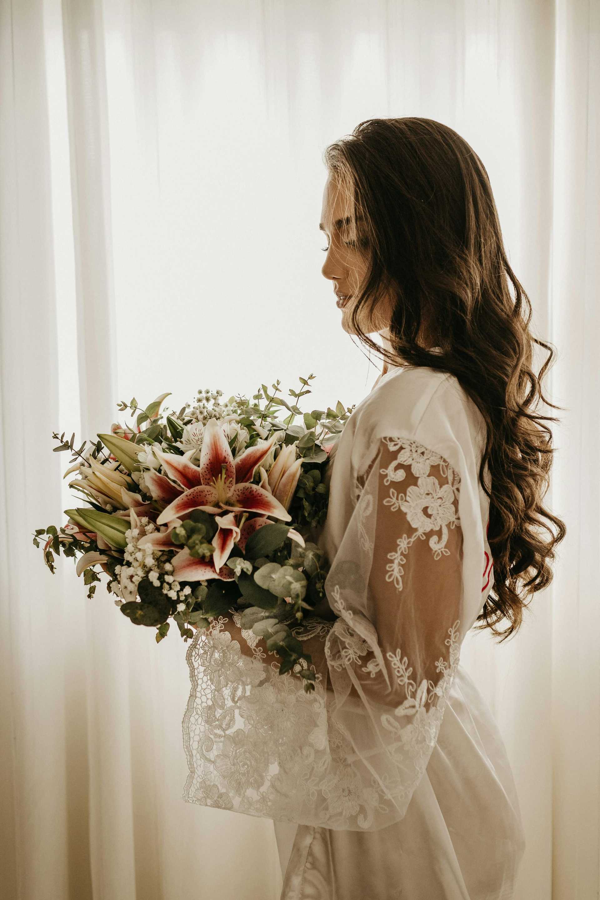 Bride in lace robe holding a bouquet with lilies, standing by a soft-lit window.